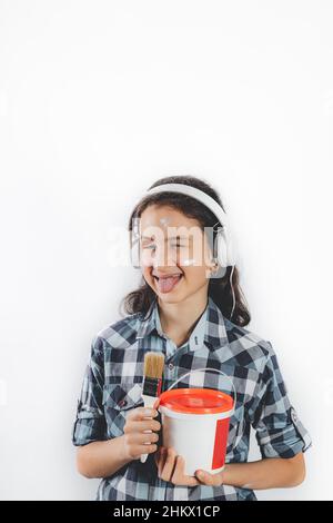 Porträt eines positiv lächelnden Teenagers. Nettes hübsches Mädchen in weißen Kopfhörern mit einem Pinsel und einer Dose Farbe auf weißem Hintergrund. Positiv Stockfoto