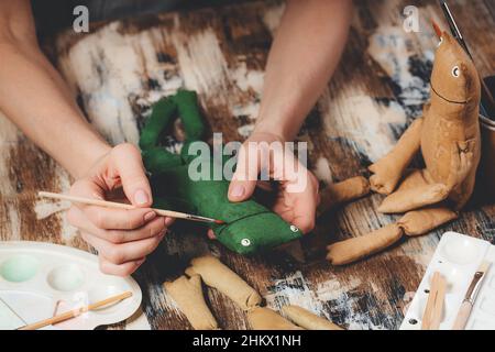 Handgefertigter weicher Puppenfrosch. Geschenke selbst gestalten. Menschliche Hände halten Frosch weiches Spielzeug. Nadelarbeitszubehör auf grauem Hintergrund. Handgemachte Einrichtung im Haus. Prozess von Stockfoto