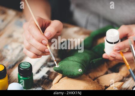 Handgefertigter weicher Puppenfrosch. Basteln von DIY-Geschenken. Menschliche Hände halten Frosch weiches Spielzeug. Nadelarbeitszubehör auf grauem Hintergrund. Handgemachte Einrichtung im Haus. Prozess o Stockfoto