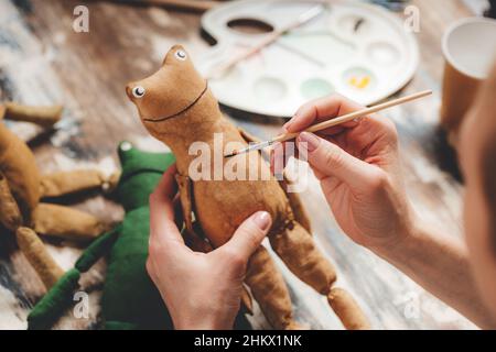 Handgefertigter weicher Puppenfrosch. Basteln von DIY-Geschenken. Menschliche Hände halten Frosch weiches Spielzeug. Nadelarbeitszubehör auf grauem Hintergrund. Handgemachte Einrichtung im Haus. Prozess o Stockfoto