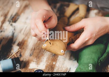 Handgefertigter weicher Puppenfrosch. Basteln von DIY-Geschenken. Menschliche Hände halten Frosch weiches Spielzeug. Nadelarbeitszubehör auf grauem Hintergrund. Handgemachte Einrichtung im Haus. Prozess o Stockfoto