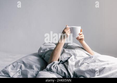 Die Hand einer Frau hält eine Tasse Kaffee, während sie im Bett liegt. Stockfoto