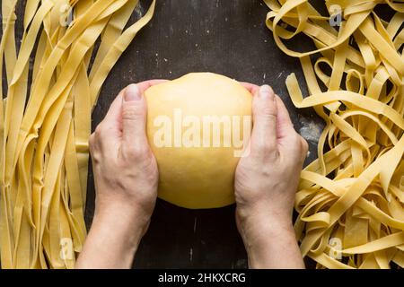 Frau Hände halten Kugel aus Teig und rohe gerade und gebogene Streifen von Nudeln auf dem hölzernen Hintergrund Stockfoto