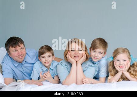 Glückliche kaukasische große Familie liegt im Schlafzimmer auf dem Bett und lächelt. In blauer Kleidung gekleidet haben Spaß zusammen. Eltern und Kinder zwei Söhne und eine süße Tochter Stockfoto