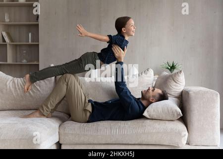 Glücklicher Vater liegt auf der Couch und hebt die Tochter so, als ob sie fliegen würde Stockfoto