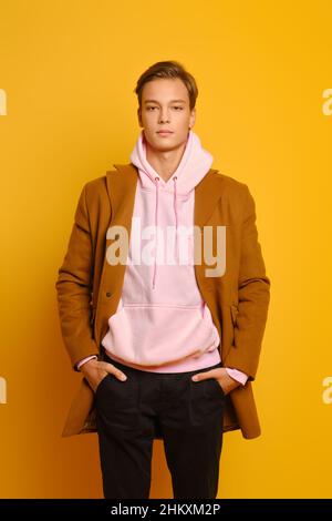 Hübscher, stylischer Mann in braunem Mantel und pinkfarbenem Kapuzenpullover mit Händen in Taschen auf gelbem Studiohintergrund Stockfoto