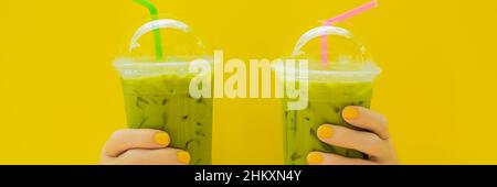 Zwei grüne Kaffee Latte mit Eis in Plastikbecher und Stroh in weiblicher Hand mit gelben Maniküre auf gelben Hintergrund. Hausgemachte Eistee Matcha Latte Tee mit Stockfoto