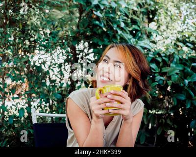 Porträt einer glücklichen, schönen erwachsenen asiatischen Frau mit einem Highlight trendige kurze Frisur Kaffee trinken, halten die gelbe Tasse, nach oben schauen, und sitt Stockfoto