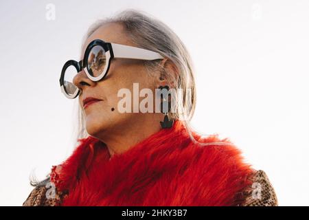 Nachdenkliche ältere Frau schaut weg, während sie im Freien steht. Stilvolle ältere Frau in farbenfroher Freizeitkleidung und Brillen. Reife Frau, Reflec Stockfoto