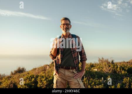 Reifer Wanderer lächelt bei Sonnenuntergang an der Kamera. Fröhlicher, reifer Wanderer, der auf einem Hügel steht und dabei einen Rucksack trägt. Abenteuerlicher männlicher Rucksacktourist enjo Stockfoto