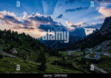 Eine kurvenreiche Straße, die zum Pordoipass führt, die Sellagruppe auf der rechten Seite, Gipfel von Plattkofel, Grohmannspitze und Langkofel in der Ferne bei Sonnenuntergang Stockfoto