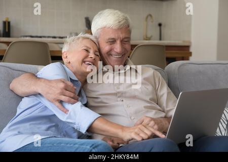 Fröhliches älteres Ehepaar mit 60s Enkeleltern, das einen Laptop auf der Couch benutzte Stockfoto