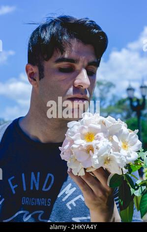 Romantischer junger argentinischer Mann in der Liebe in einem Park, der einen Strauß weißer Blumen, vertikales Bild, Liebe und Lifestyle-Konzept riecht. Stockfoto