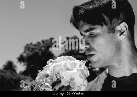 Dramatischer und romantischer junger kaukasischer argentinischer Mann in Schwarz und Weiß mit geschlossenen Augen, die eine Blume im Park riechen, mit dem Himmel im Hintergrund und Stockfoto