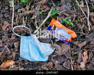 Eine ausrangierte Gesichtsmaske aus blauem Kunststoff und eine Schokoriegel-Verpackung gegen heruntergefallene Blätter in einem ländlichen Ort im Winter in Großbritannien. Stockfoto