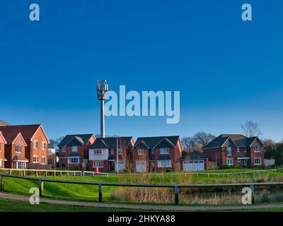 Ein hoher Handymast neben modernen Wohnhäusern mit einer Rasenfläche und einem Teich im Vordergrund. Aufgenommen an einem sonnigen Tag im Winter. Stockfoto