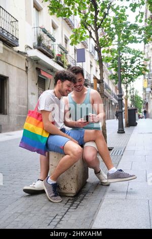 Schwules Paar mit dem Smartphone und lachend auf der Straße sitzen. Stockfoto