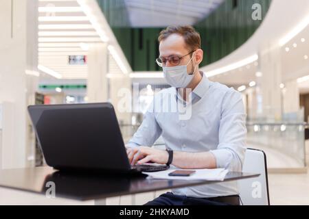 Angestellter mit Gesichtsmaske, Mann, der an einem öffentlichen Ort an einem Laptop arbeitet Stockfoto