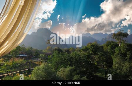 Fensterblick über weißen Vorhang auf grünes tropisches Tal und Karstberge, Sonnenstrahlen brechen Morgenwolken - Vang Vieng, Laos Stockfoto