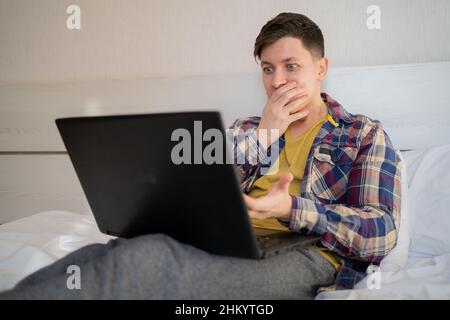 Ein Fernarbeiter mit einem Laptop sitzt tagsüber in legerer Kleidung zu Hause auf dem Bett, bedeckt seinen Mund mit der Hand, schockiert und ängstlich vor einer MIS Stockfoto