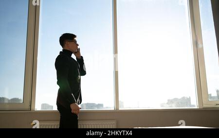 Ein männlicher Geschäftsmann steht im Büro in der Nähe des Panoramafensters und telefoniert mit seinem Mobiltelefon. Anwendungen und Lösungen für digitale Technologien Stockfoto