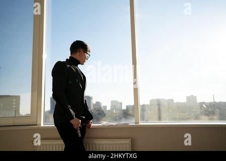Seitenansicht eines jungen erfolgreichen Geschäftsmannes, der in einem Büro steht und durch ein durchgehendes Fenster auf das Stadtbild blickt, träumend oder entspannend, Stockfoto
