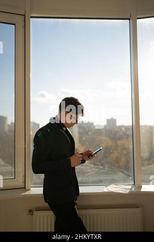 Ein junger Geschäftsmann in einem schwarzen Anzug steht neben dem Panoramafenster im Büro und nutzt sein Smartphone, liest die Nachrichten online. Stockfoto