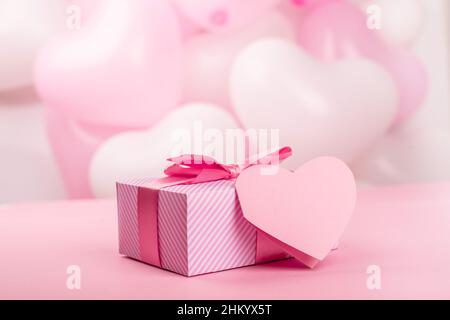 Valentinstaggeschenk in einer Schachtel, die in Streifenpapier verpackt ist und mit Seidenbandbogen und herzförmiger Grußkarte auf pinkfarbenem Ballonhintergrund mit Cop gefesselt ist Stockfoto