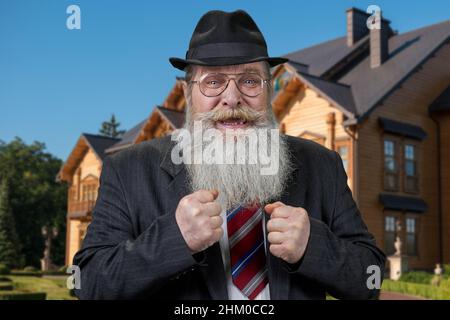 Aufgeregter, fröhlicher alter Mann, der mit geballten Fäusten im Freien steht. Schönes Holzhaus im Hintergrund. Stockfoto