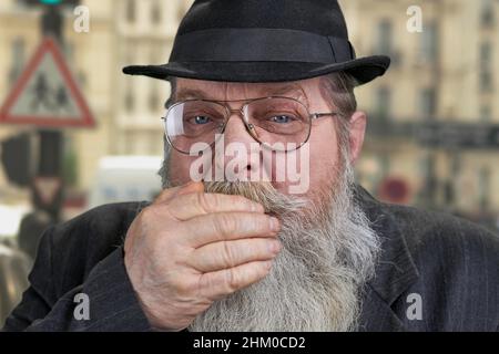 Alter bärtiger Mann, der den Mund mit der Hand aus der Nähe bedeckt. Unscharfer Hintergrund. Stockfoto
