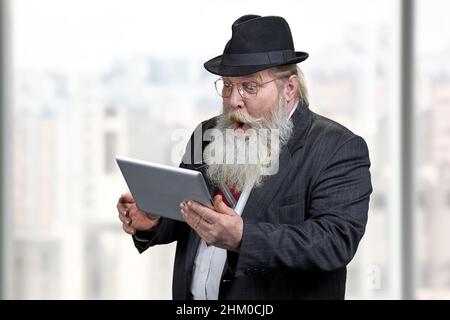 Lustig schockiert Senior Geschäftsmann mit Tablet-pc. Verschwommener Hintergrund des Bürofensters. Stockfoto