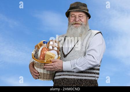 Leitender Mann hält Korb mit Milchprodukten und blickt auf die Kamera auf blauem Himmel Hintergrund. Bio-Lebensmittelproduktion. Stockfoto