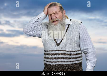 Deprimierter alter bärtiger Mann, der seinen Kopf auf bewölktem Himmel berührte. Menschliche negative Ausdrücke. Stockfoto