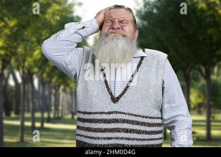 Der alte bärtige Mann trägt einen legeren Pullover, der den Kopf in den Händen hält und Kopfschmerzen oder Stress empspürt. Verzweifelter pensionierter Mann im Freien. Stockfoto