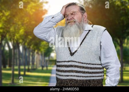 Ein alter gestresster Mann berührte seinen Kopf und hielt die Augen geschlossen, während er im Freien stand. Stress, Depression und Problemkonzept. Stockfoto