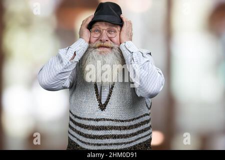 Fröhlicher, staunender alter Mann mit grauem Bart, der die Hände am Kopf hält und einen Schock ausdrückt. Abstrakter Bokeh-Hintergrund. Stockfoto