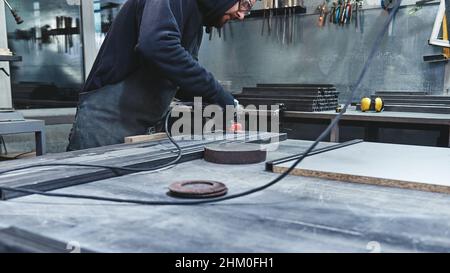 Beschnittenes Bild eines Arbeiters, der spezielle Schutzhandschuhe und eine Brille trägt und mit dem Schleifer ein Stück Metallrahmen polieren kann, das auf den Arbeitstisch gedrückt wird. Mann mit Handschleifer. Horizontale Aufnahme Stockfoto