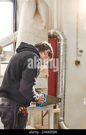 Porträt eines ausgebildeten Zimmermanns, der in seiner Schreinerei arbeitet. Holzarbeiter schneiden Holzbrett mit Handsäge für maßgeschneiderte Möbel. Geübter Handwerker sägt Holzbrett auf Tisch mit tragbarer Armsäge. Vertikale Aufnahme Stockfoto