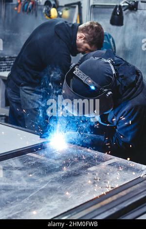 Industriearbeiter mit Helm und Handschuhen beim Schweißen von Stahlkonstruktionen in der Fabrik. Mitarbeiter im Hintergrund. Vertikale Aufnahme Stockfoto