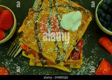 Quadratische belgische Waffeln mit Beeren und Puderzucker auf einem schwarzen Teller. Leckere süße, zuckerhaltige Waffeln. Geschmolzene Schokolade und Schlagsahne auf Waffeln. Oben Stockfoto