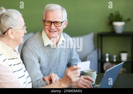 Glücklicher älterer Mann mit einem Getränk, der seine Frau mit Kreditkarte ansieht, während er online Lebensmittel zum Mitnehmen bestellt Stockfoto