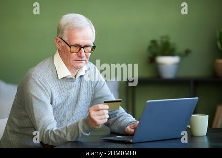 Moderner älterer Mann in blauem Pullover, der vor dem Laptop sitzt und die Nummer der Kreditkarte eingibt, während er online bezahlt Stockfoto