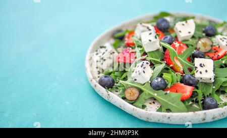 Salat mit Rucola, Schafskäse und Beeren - Erdbeere, Heidelbeere, im Handwerk. Sommersalat Idee und Rezept für gesundes vegetarisches Mittagessen, Abendessen. Close Up. Kopieren Sie Platz. Bunte blauen Hintergrund. Stockfoto