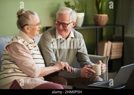 Glücklicher Rentner, Ehemann und Ehefrau in legerer Kleidung, die im Wohnzimmer auf der Couch vor dem Laptop saßen und sich unterhalten haben Stockfoto