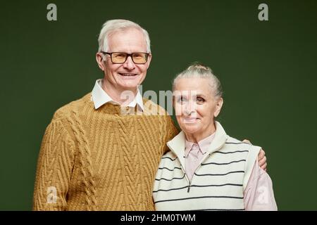 Porträt eines glücklichen älteren Mannes in braunem Pullover, der seine Frau in gestreifter Freizeitkleidung auf dunkelgrünem Hintergrund umarmt Stockfoto