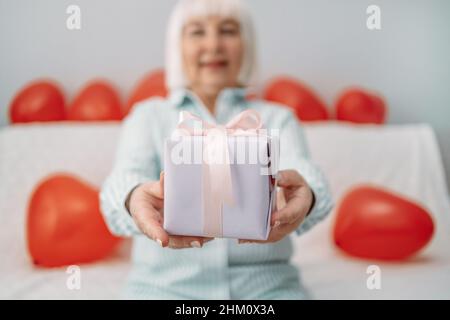 Lächelnde, 50 Jahre alte blonde Frau, die am 14. februar auf einem Sofa auf grauem Hintergrund zu Hause einen Geschenkkarton auf der Kamera gibt Stockfoto