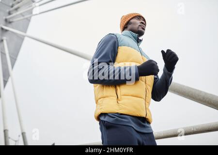 Aktiver junger afroamerikanischer Mann in Sportkleidung und schwarzen Handschuhen, der morgens in der städtischen Umgebung läuft Stockfoto