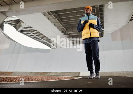 Junger, aktiver afroamerikanischer Mann in warmer Sportbekleidung läuft auf der Joggingstrecke während des sportlichen Trainings im Freien Stockfoto