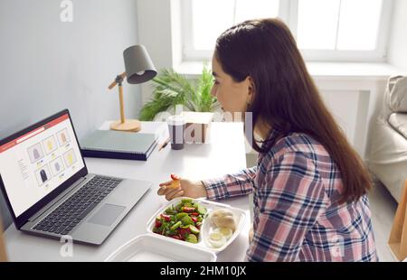 Frau isst beim Lieferservice bestellte Lebensmittel, während sie in ihrem Heimbüro vor dem Laptop sitzt. Stockfoto