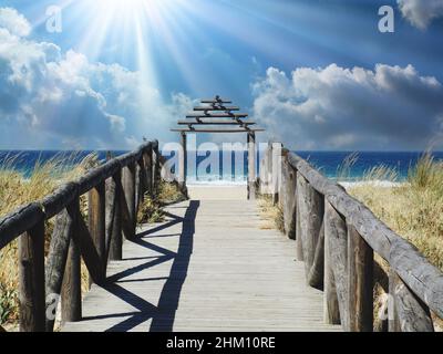 Blick auf hölzernen Pier mit Geländer und Tor über Sanddünen zum blauen Ozean und Himmel mit Sonnenstern und Cumuluswolken (Fokus auf Tor) Stockfoto